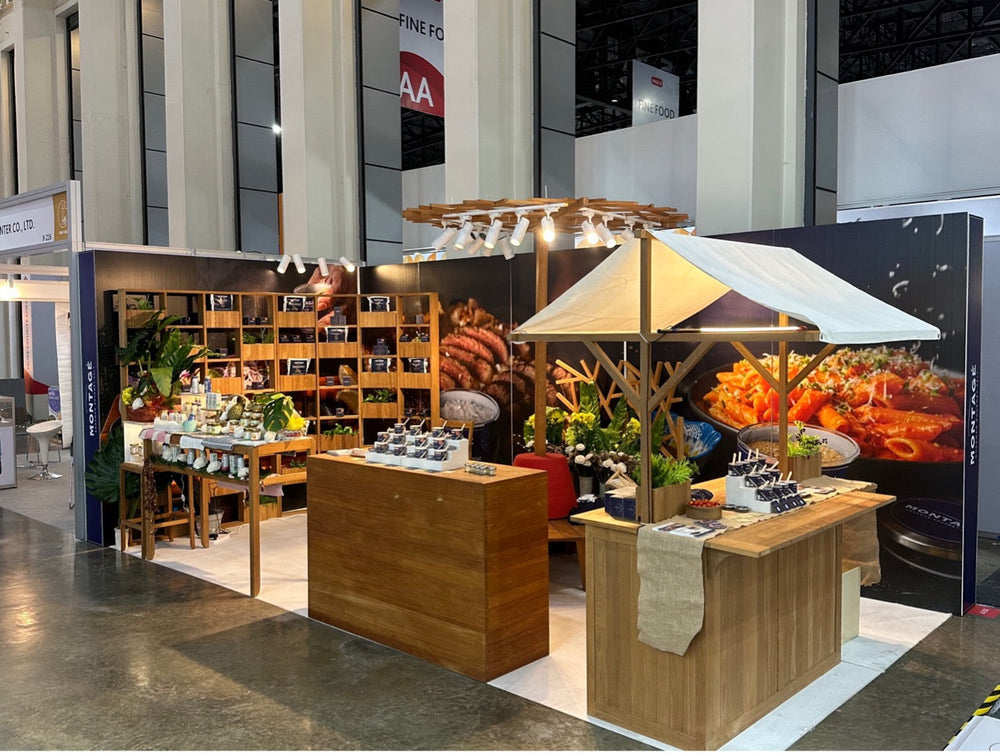 A premium display of luxury salt and salt flakes at the Montagé booth during THAIFEX. The booth showcases bulk gourmet food salt in stylish packaging arranged on artisanal wooden displays and a rustic market cart.