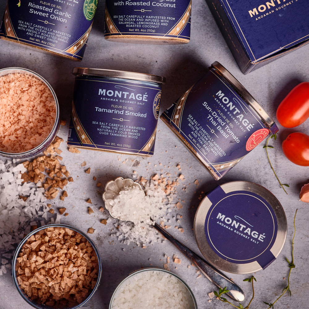 Montagé finishing salt tins — Tamarind Smoked, Sun-Dried Tomato Thai Basil, and Roasted Garlic Sweet Onion — arranged with loose salt and fresh tomatoes
