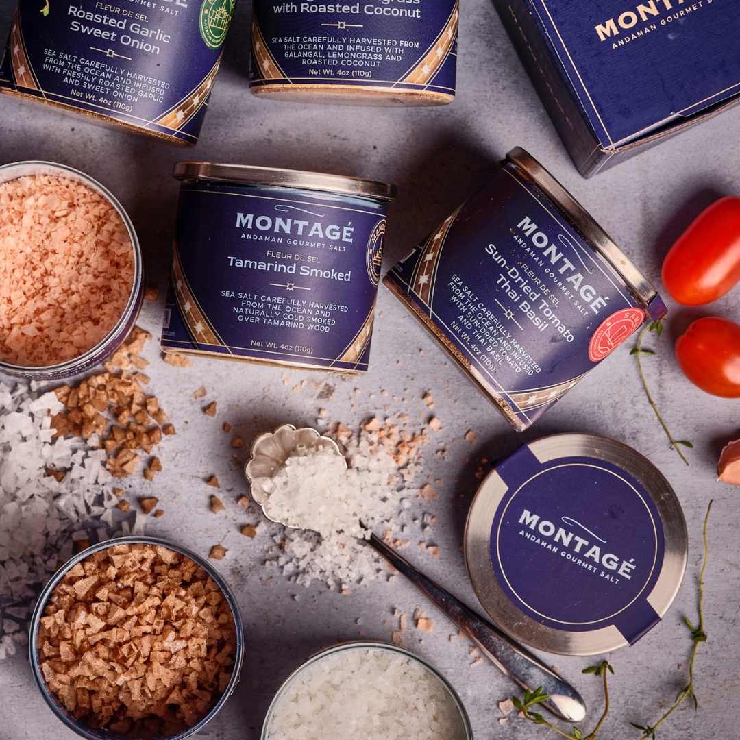 Montagé finishing salt tins — Tamarind Smoked, Sun-Dried Tomato Thai Basil, and Roasted Garlic Sweet Onion — arranged with loose salt and fresh tomatoes