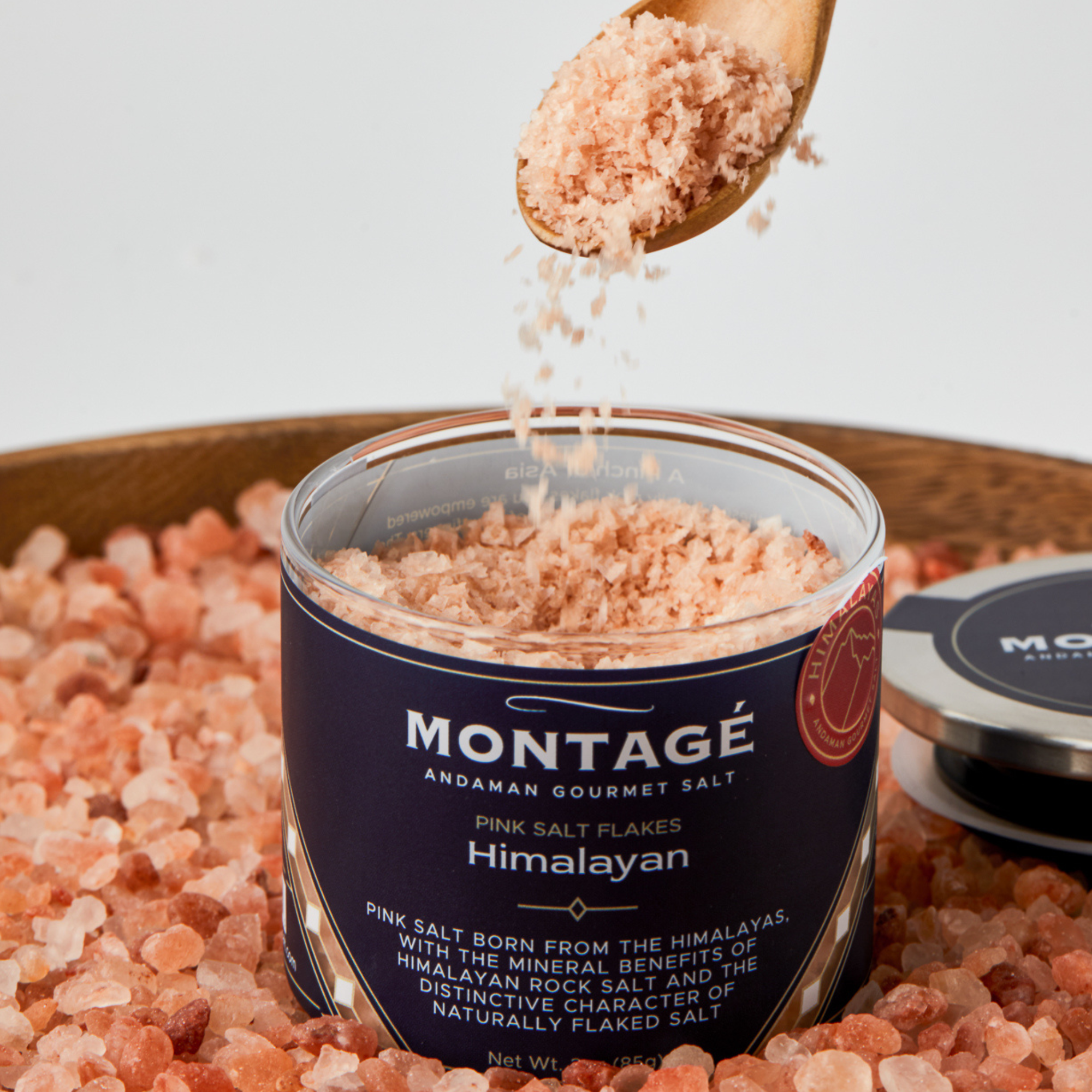 Flake pink himalayan salt poured from a wooden spoon into a glass jar with a blue label