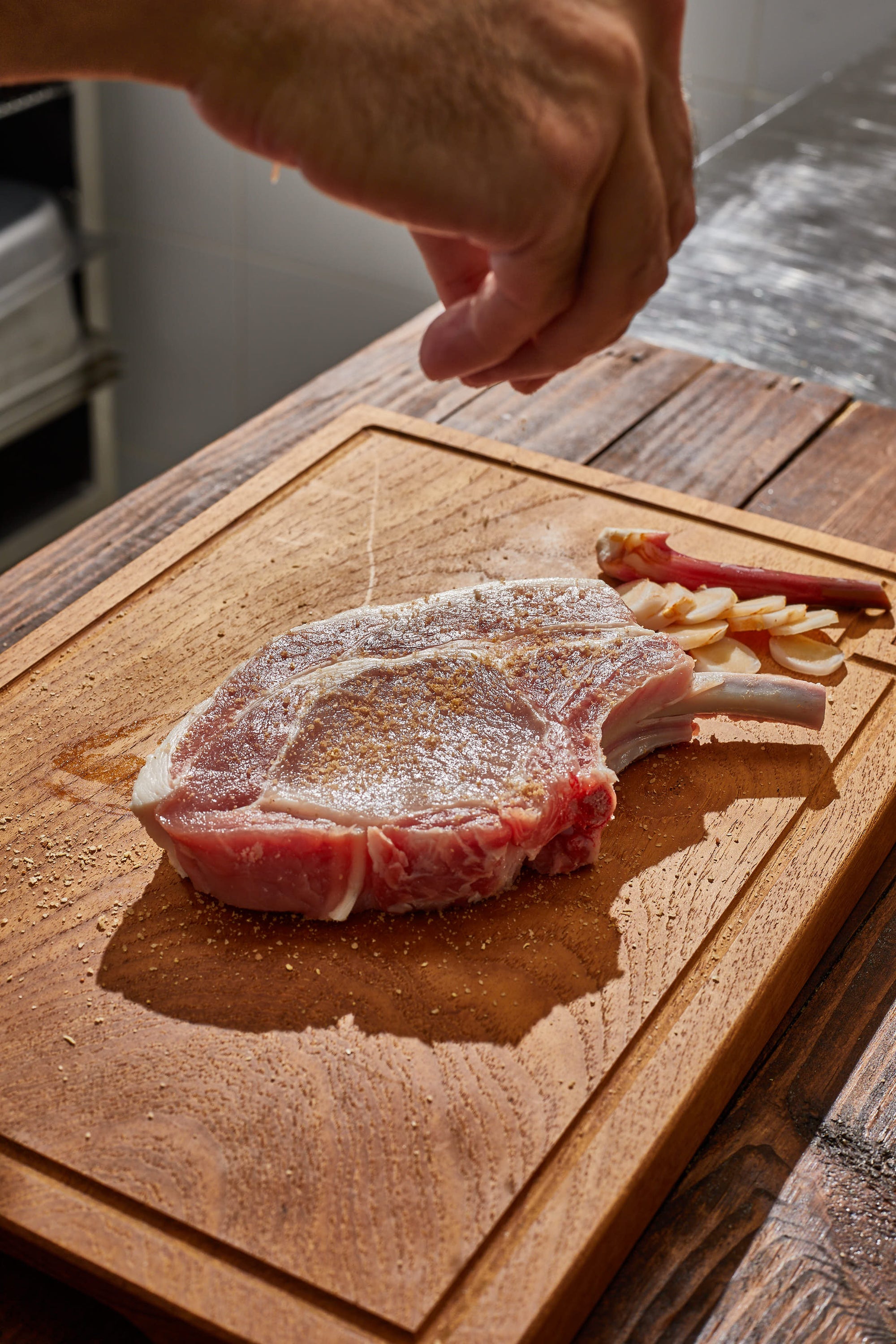 Raw pork chop on a wooden cutting board with a hand sprinkling seasoning.