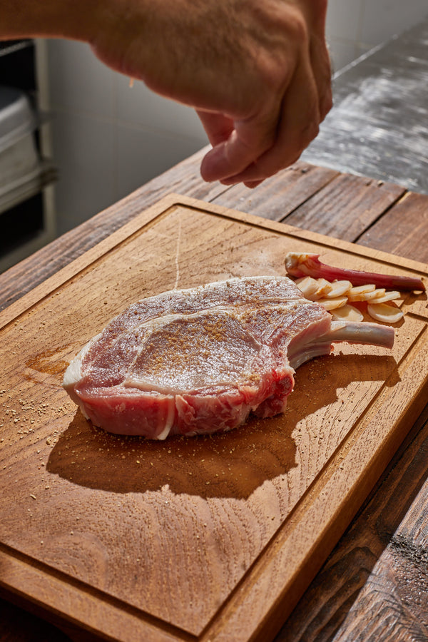 Raw pork chop on a wooden cutting board with a hand sprinkling seasoning.