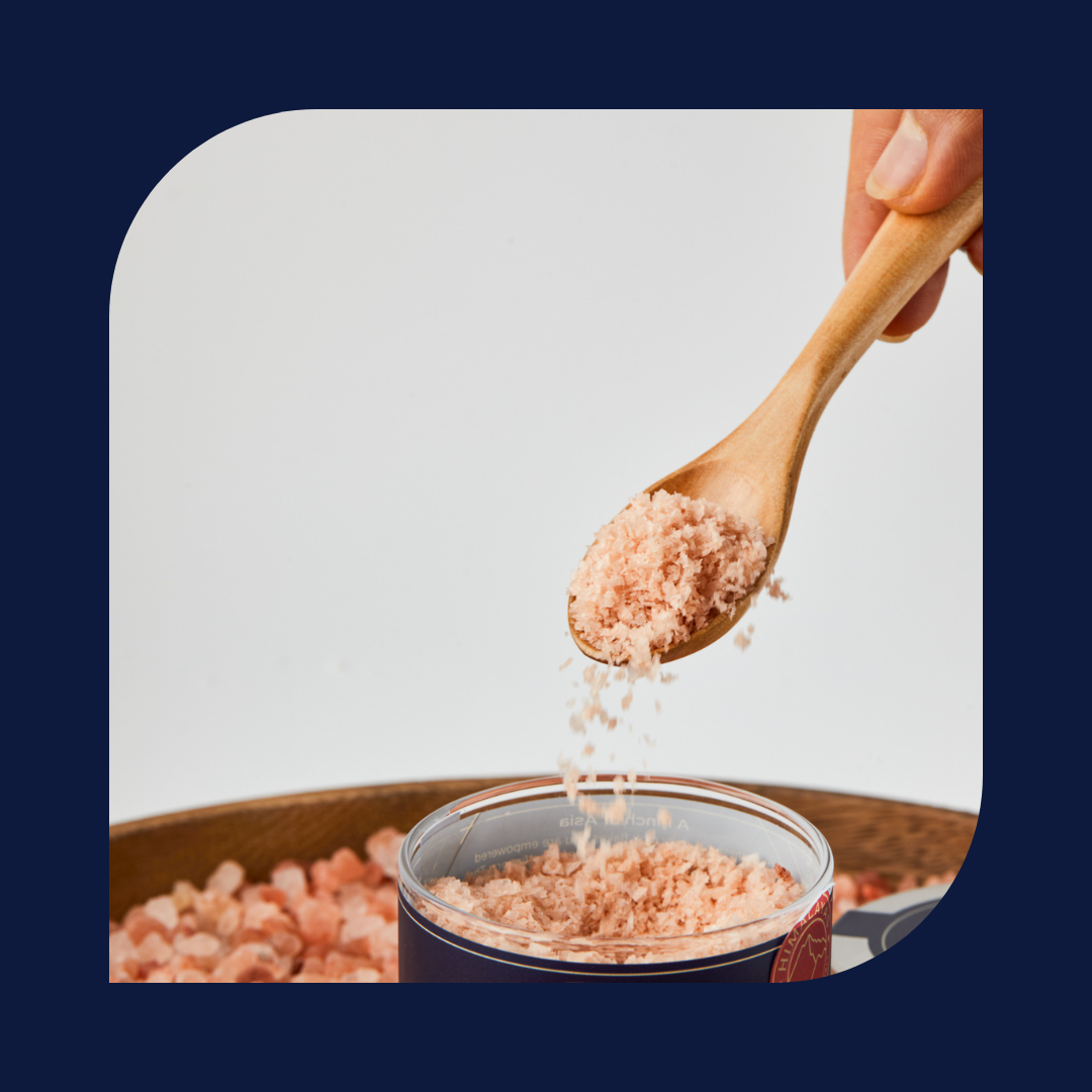Wooden spoon pouring pink Himalayan salt flakes into a glass container with a blue border.