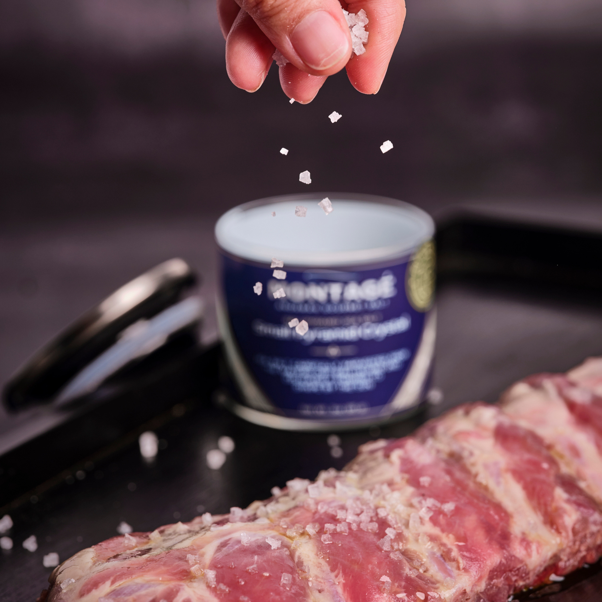 Hand sprinkling coarse salt over steak meat with a container of coarse kosher salt in the background.