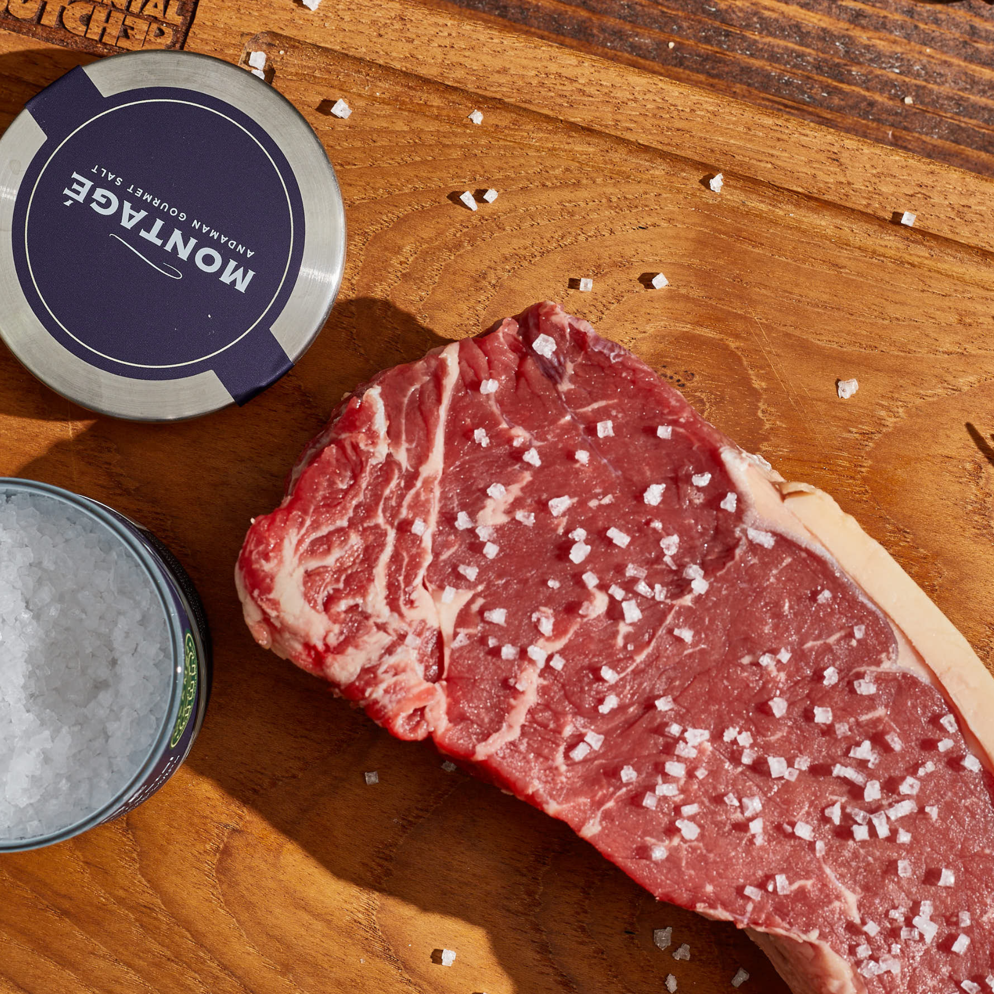 Raw steak garnished with coarse salt on a wooden cutting board, featuring a Montage container.