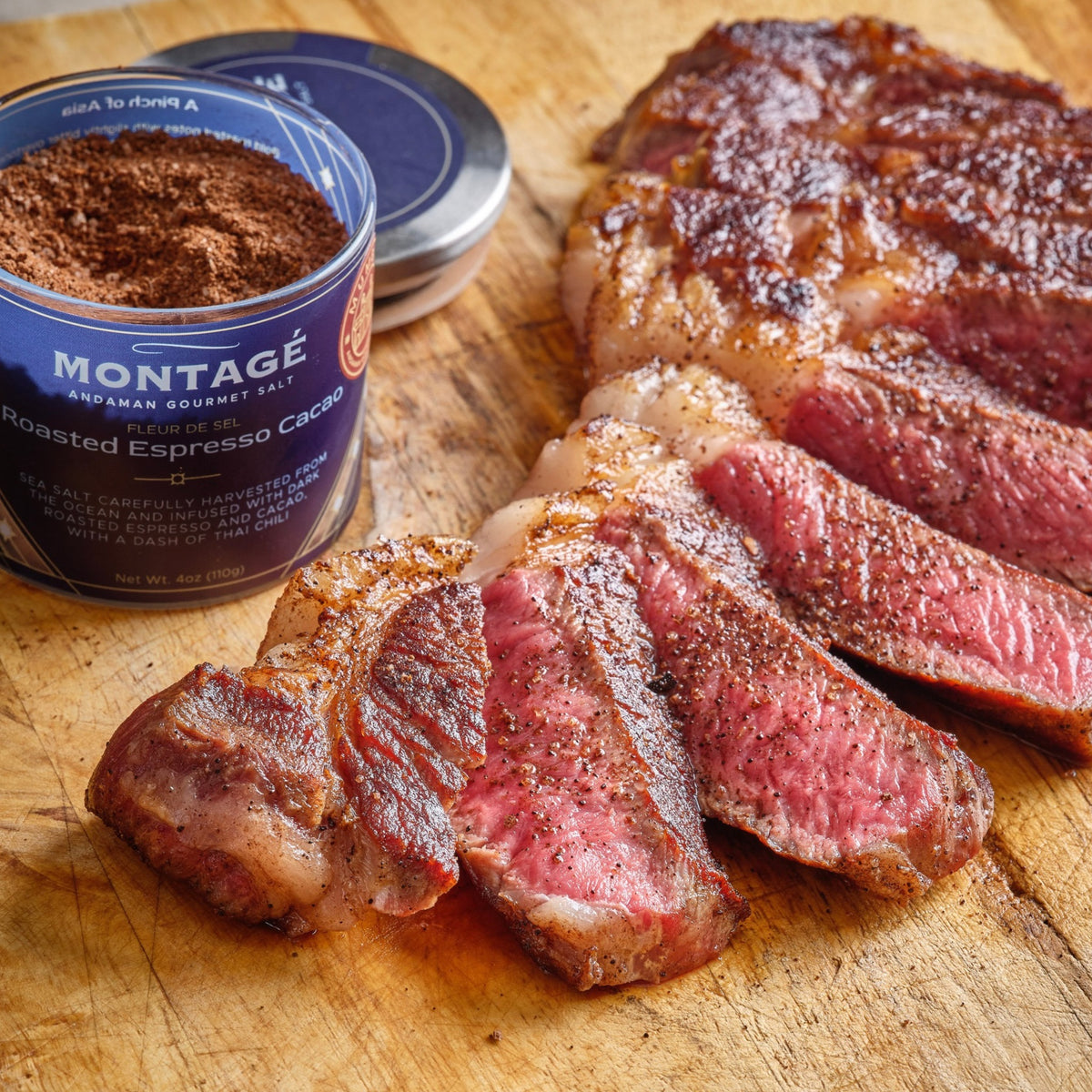 Sliced seared steak on a cutting board beside an open jar of Montagé Roasted Espresso Cacao finishing salt