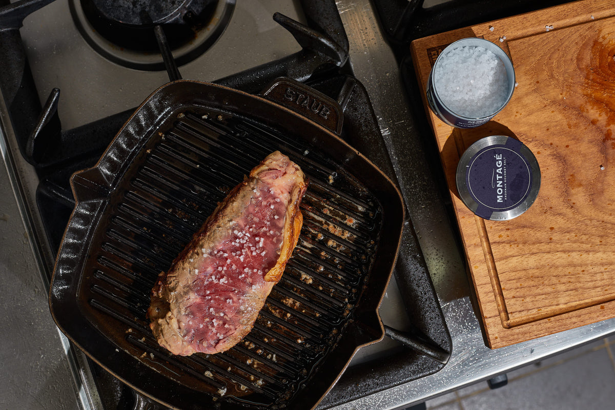 Strip steak pan-searing in a Staub cast iron grill pan next to Montagé Kusamba salt