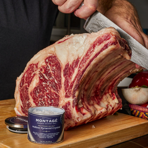 Trimming a raw bone-in prime rib roast next to a jar of Montagé Tejakula hollow pyramid sea salt