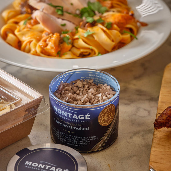 A glass jar of Montagé Longan Smoked Sea Salt, with a blue label, placed on a kitchen counter alongside a canopy of pasta and a piece of cooked chicken.